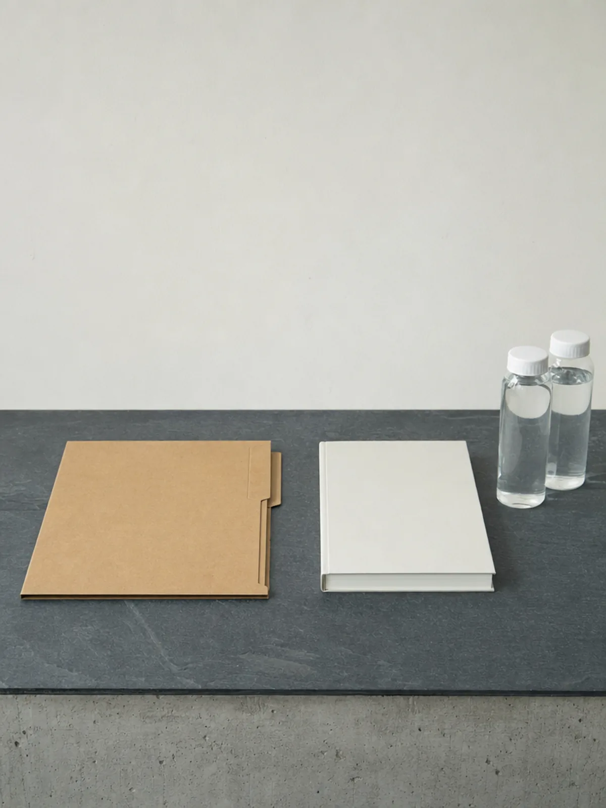 A dark slate worktop carrying one closed kraft folder, a plain bound notebook and two capped clear sample bottles against a pale wall.