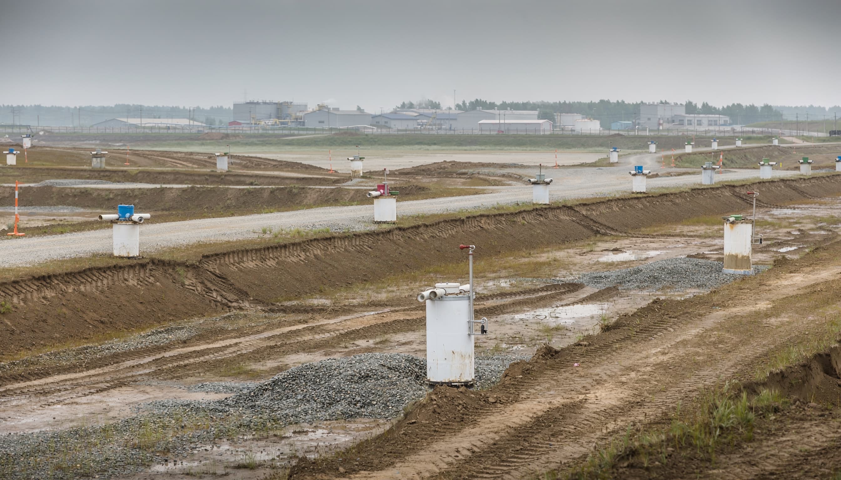 Wide horizontal panorama of a remediation site at scale: containment berms, monitoring wells, distant facility at horizon.