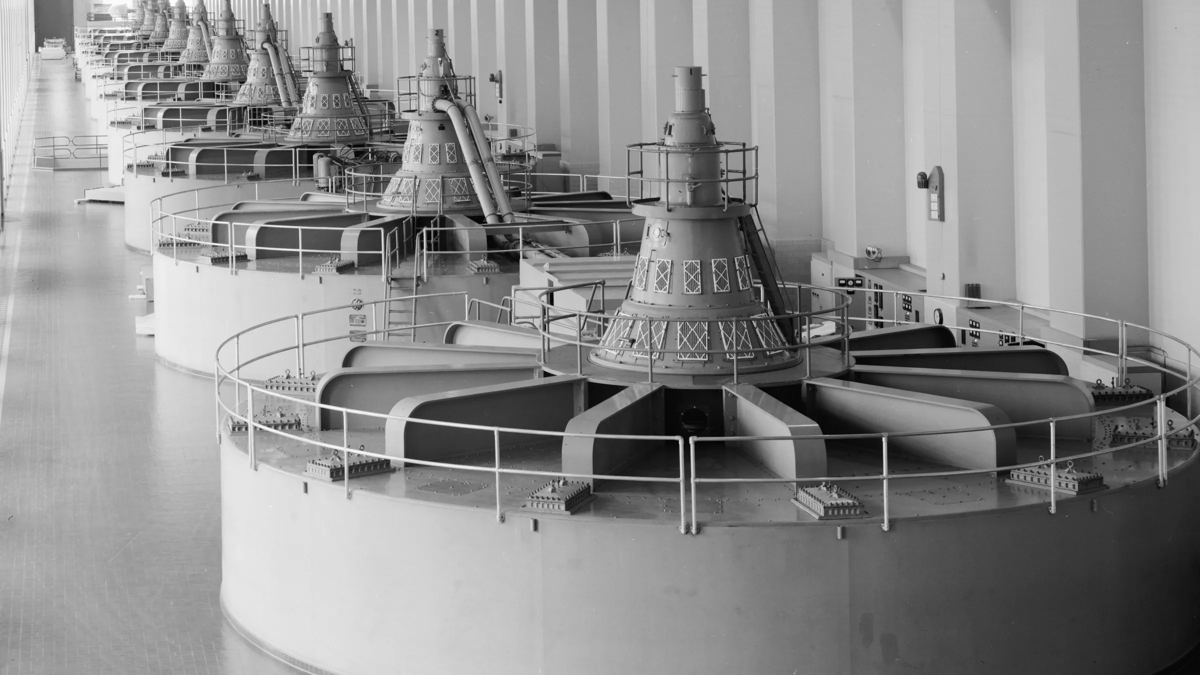 Black-and-white photograph of the generator floor of Bonneville Dam's first powerhouse, a row of round generator housings receding down a long white hall, 1987.