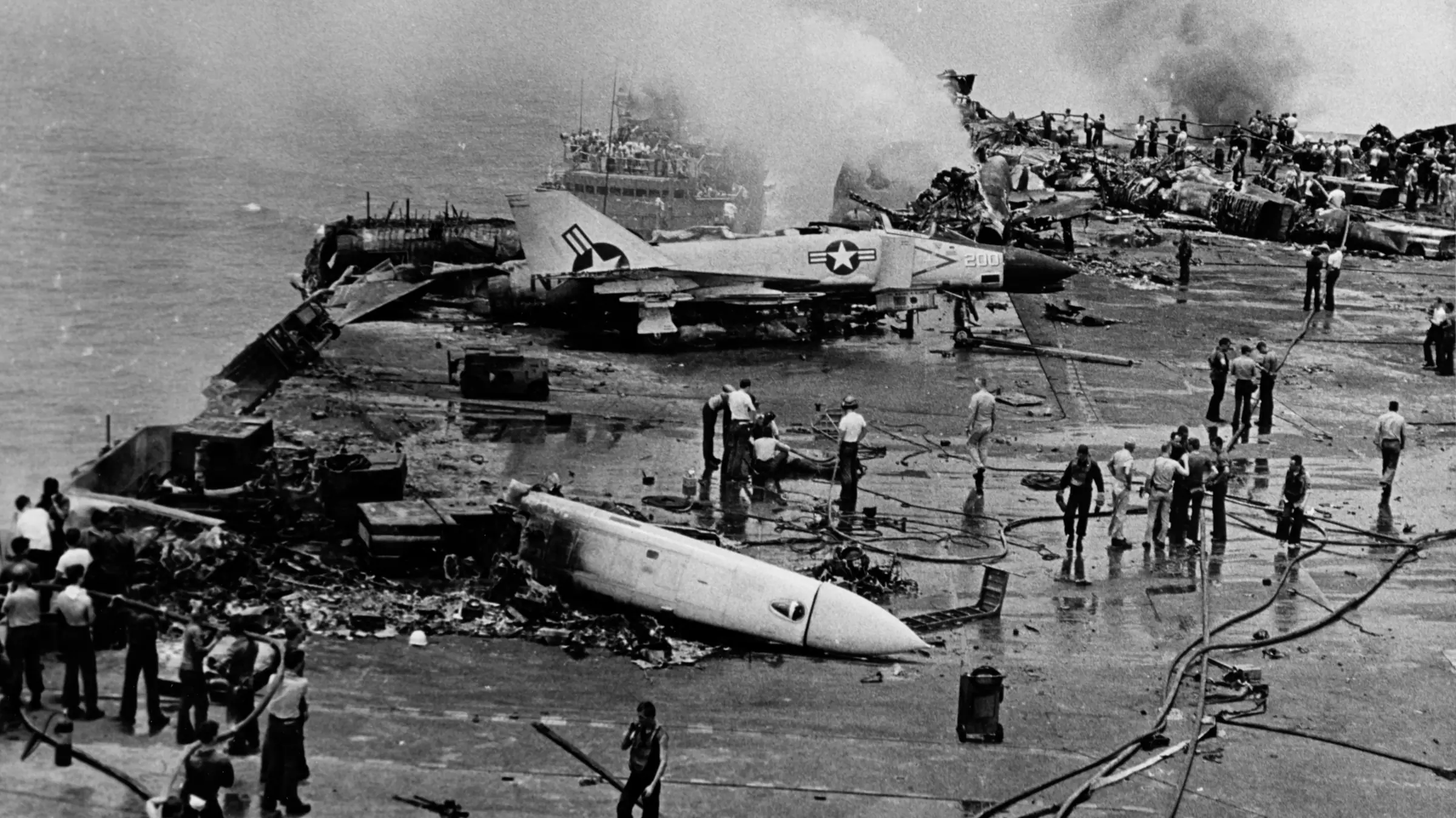 Black-and-white photograph of sailors with hoses on the charred flight deck of USS Forrestal, a damaged jet and wreckage around them and smoke rising, July 29, 1967.
